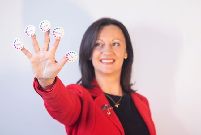 Top-Five-Marketing-Ideas Yvonne is show holding our her hand and on each finger is a sticker with the Essential Print Services' logo. Yvonne is smiling while she looks at her hand.