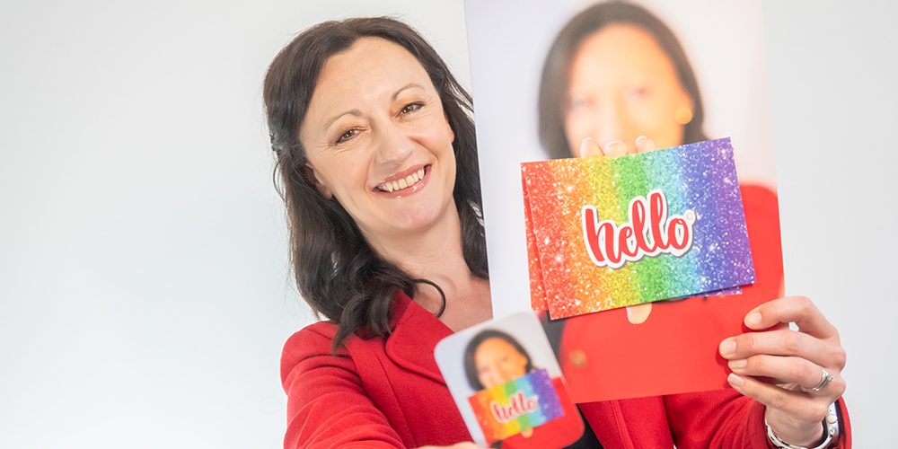 Yvonne Gorman at Derby Essential Print Services Free Artwork Check Yvonne is shown holding her business card and a brochure with the word 'Hello' on the front. Yvonne is smiling.