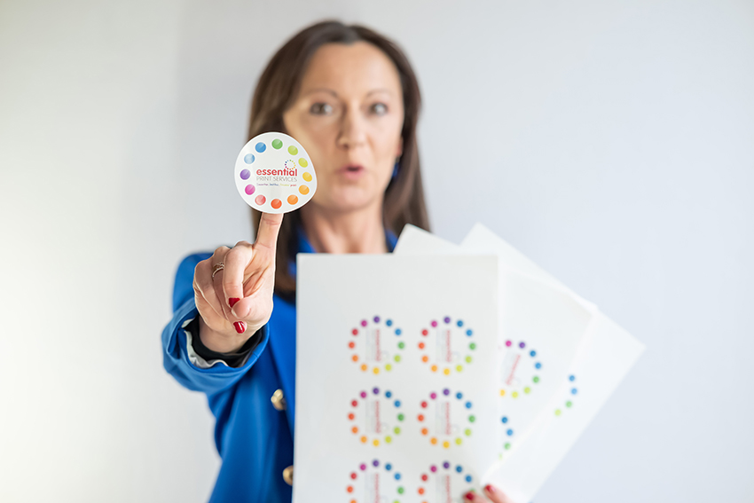 Yvonne in a bright blue blazer holds up a round, colorful sticker from Essential Print Services on her fingertip, with a surprised expression. She also holds several sheets of identical stickers in her other hand. The sticker design features a ring of multicoloured dots surrounding the company logo. The background is light and uncluttered.