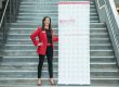 Yvonne wearing a red blazer, black outfit, and patterned high heels stands confidently next to a branded roller banner for Essential Print Services. The banner displays the company’s name, slogan, website, and a repeating circular logo pattern. They are positioned on a large indoor staircase with metal railings.