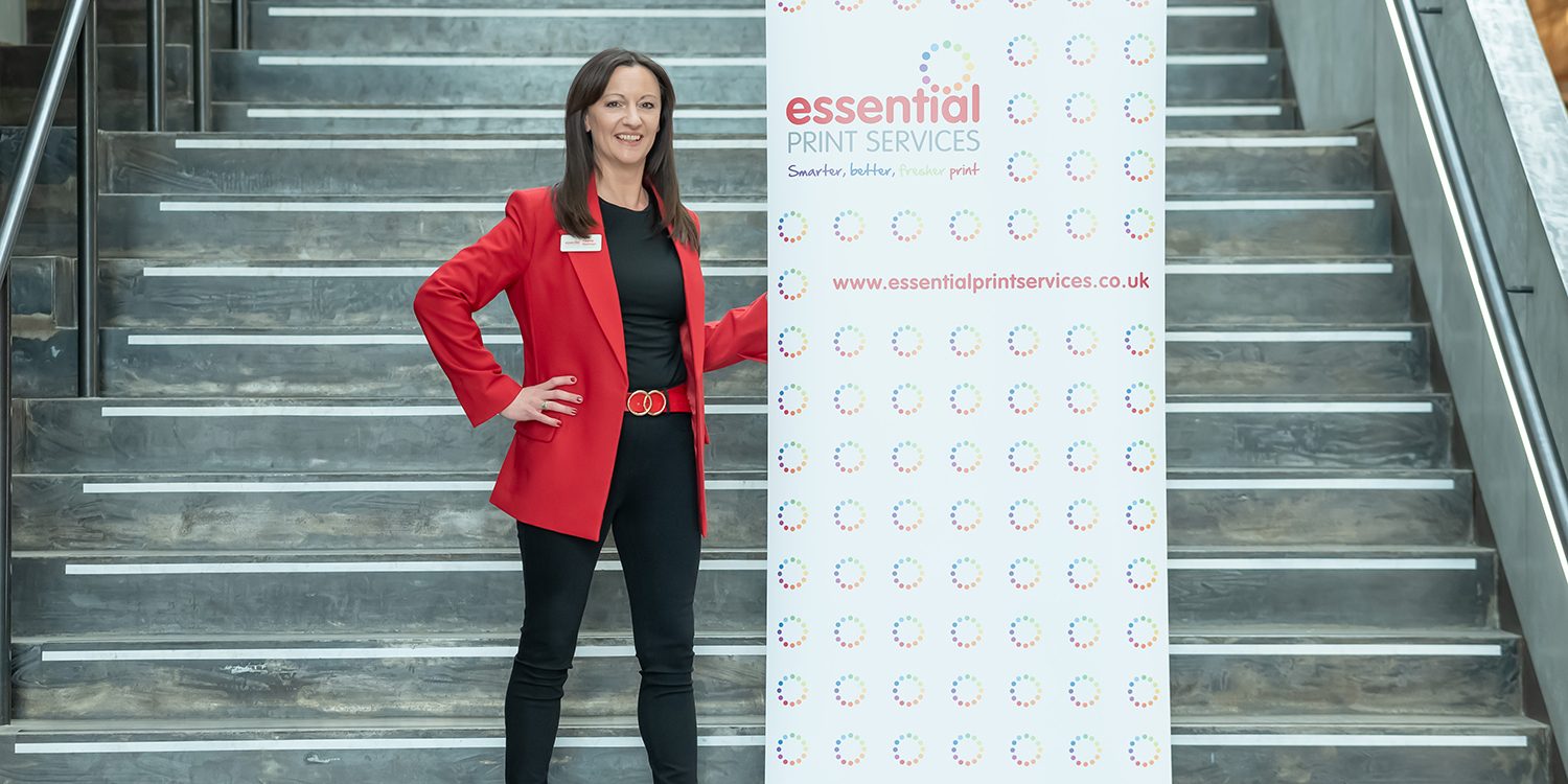 Yvonne wearing a red blazer, black outfit, and patterned high heels stands confidently next to a branded roller banner for Essential Print Services. The banner displays the company’s name, slogan, website, and a repeating circular logo pattern. They are positioned on a large indoor staircase with metal railings.