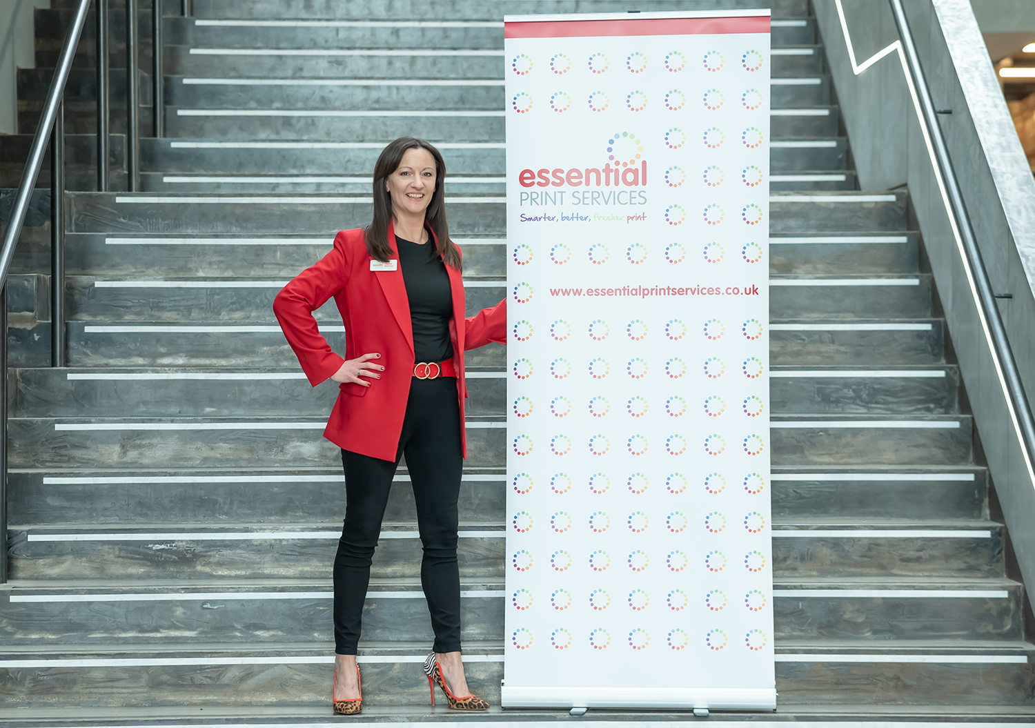 Yvonne wearing a red blazer, black outfit, and patterned high heels stands confidently next to a branded roller banner for Essential Print Services. The banner displays the company’s name, slogan, website, and a repeating circular logo pattern. They are positioned on a large indoor staircase with metal railings.
