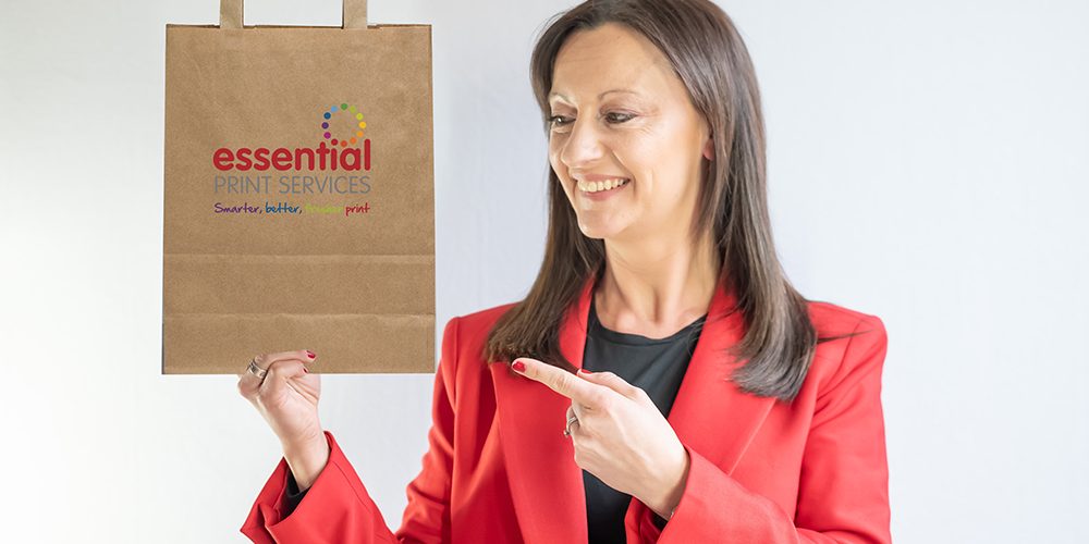 Printed-Paper-Bags Yvonne is holding a paper bag which is brown and has the Essential Print Services' logo on it. Yvonne is wearing a red blazer.