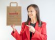 Yvonne is holding a paper bag which is brown and has the Essential Print Services' logo on it. Yvonne is wearing a red blazer.