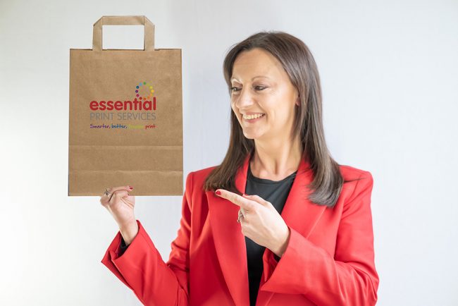 Yvonne is holding a paper bag which is brown and has the Essential Print Services' logo on it. Yvonne is wearing a red blazer.